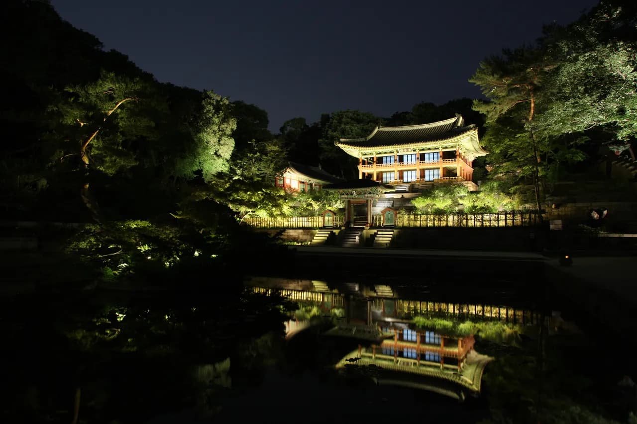 A lit Korean palace pavilion reflected in a still garden pond at night