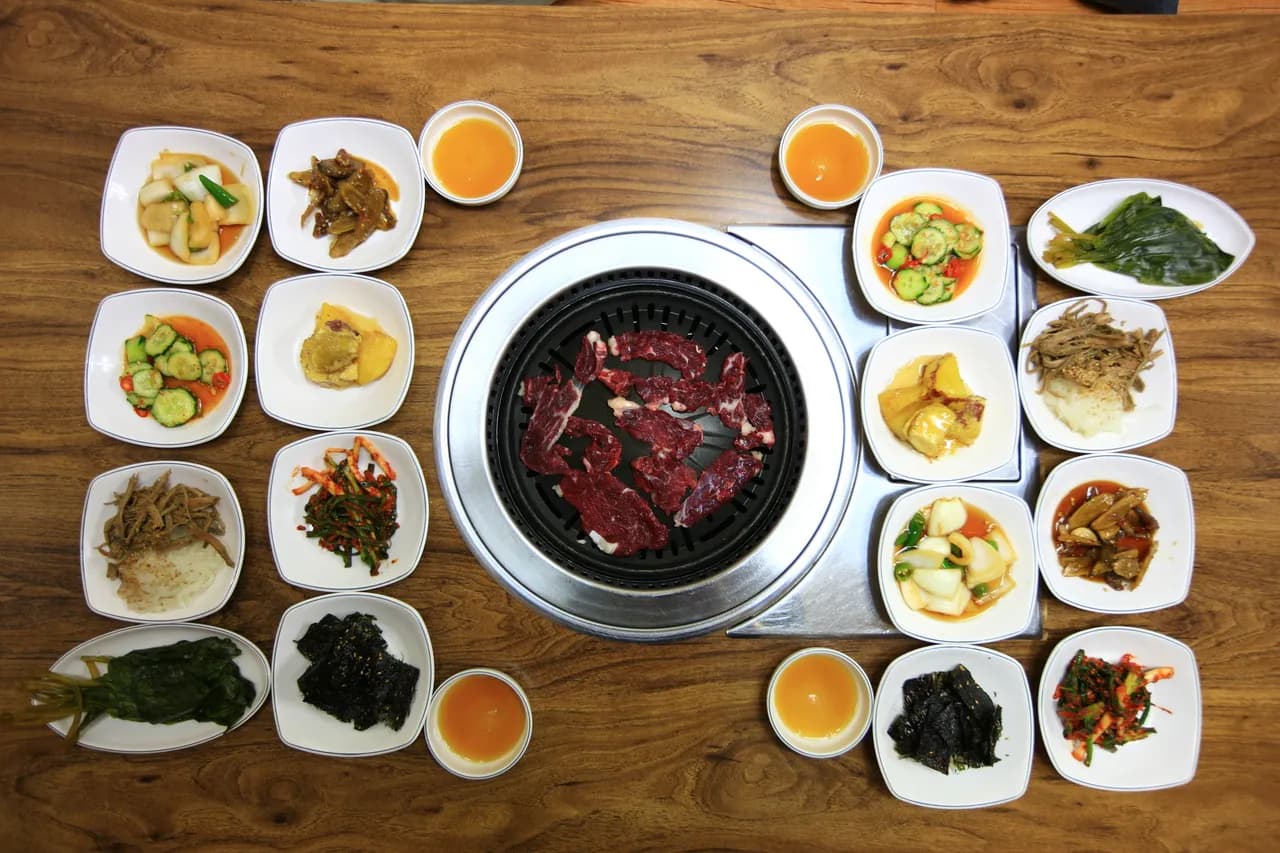 A Korean charcoal BBQ grill with raw beef in the center, surrounded by an array of banchan side dishes