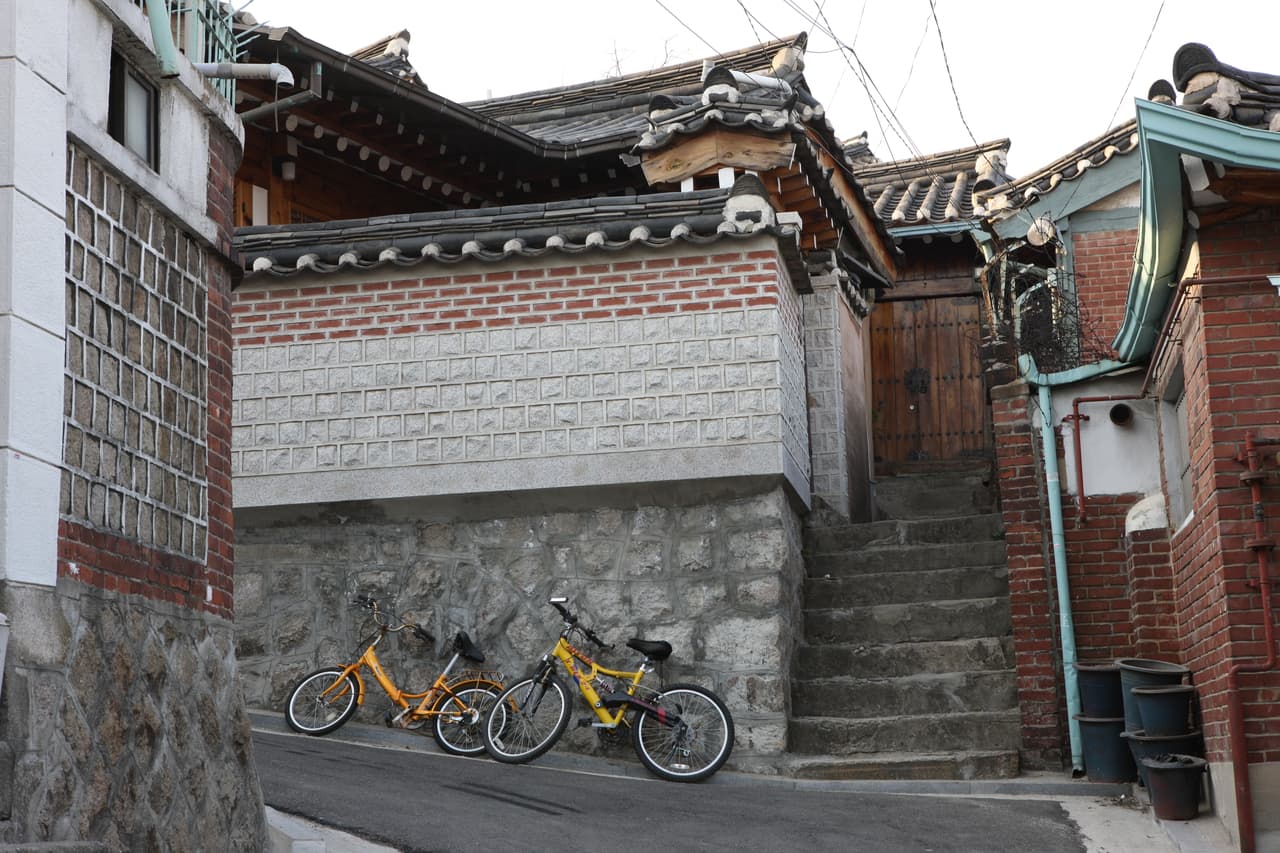 A quiet hanok corner with a stone-and-brick wall, a closed wooden gate, and parked bicycles
