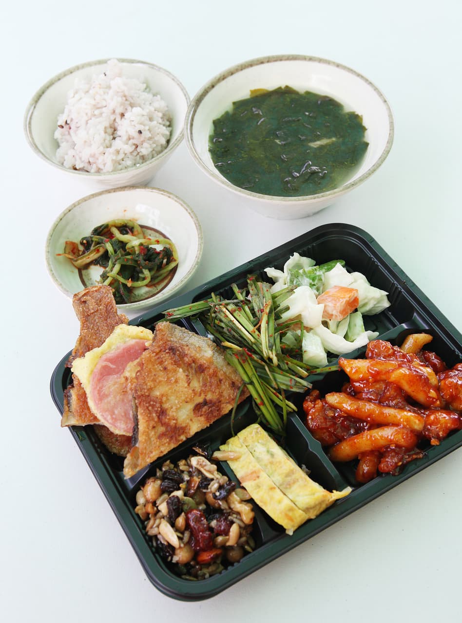 A Korean lunchbox tray filled with jeon, tteokbokki, and side dishes beside rice and soup