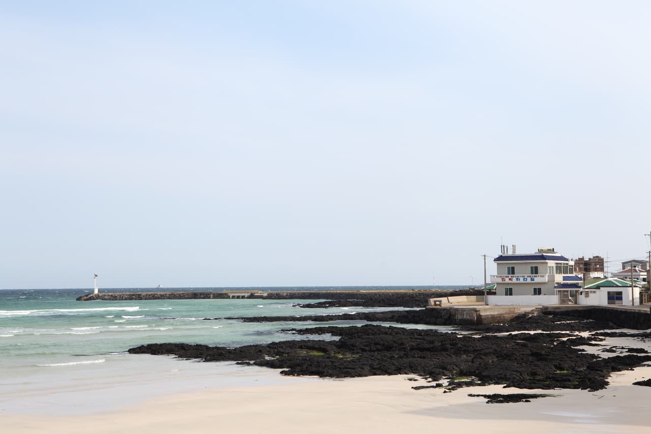 A quiet Jeju beach with white sand, emerald water, volcanic rocks, and a small lighthouse