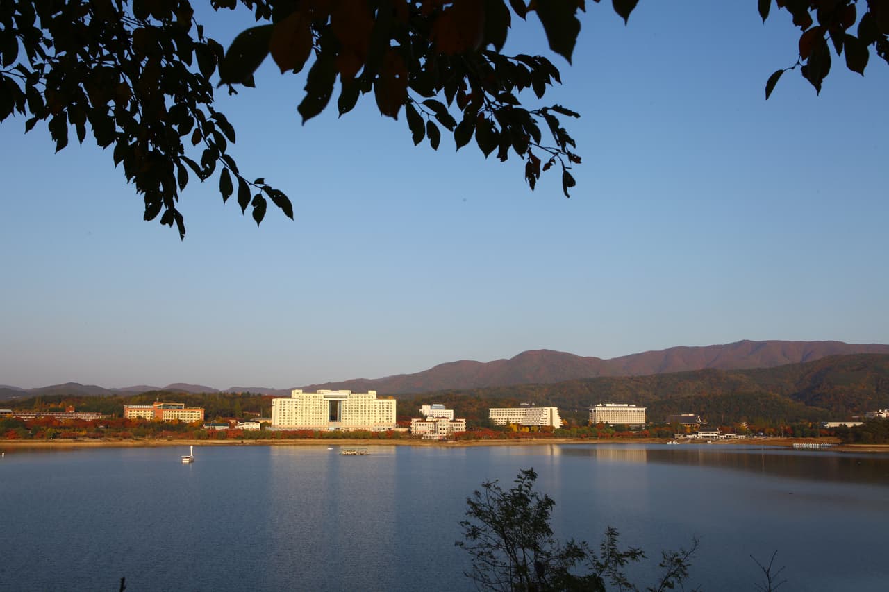 A calm lake with low resort buildings and wooded hills under a clear autumn sky
