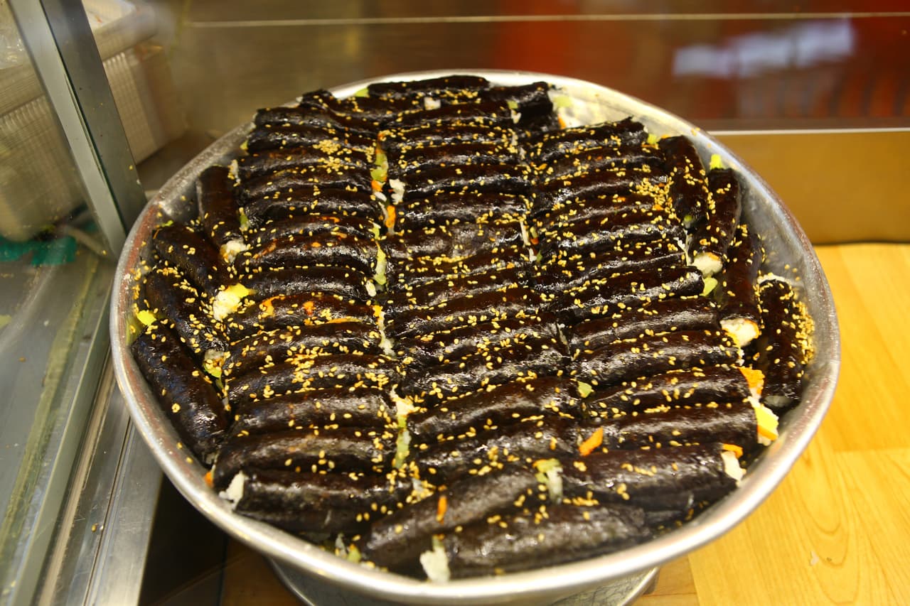 A large tray of mayak gimbap — small seaweed rice rolls topped with sesame seeds — at a market stall