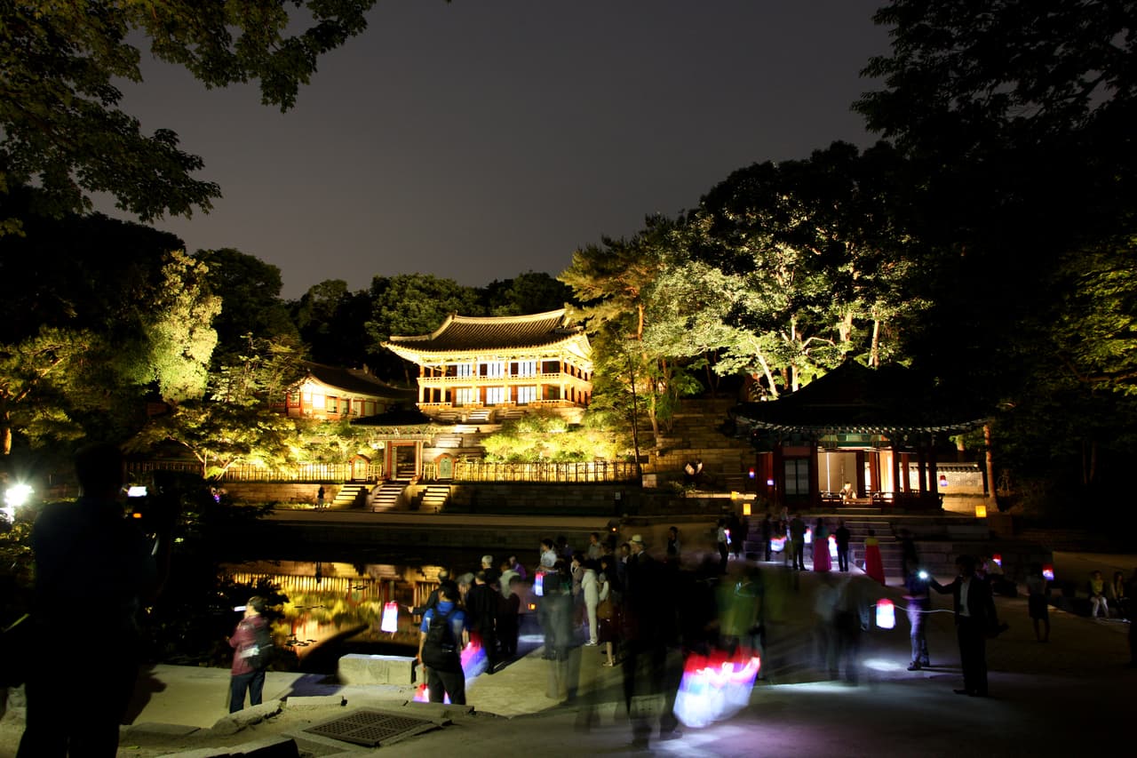 Visitors carrying paper lanterns on a guided night walk through a lit palace garden