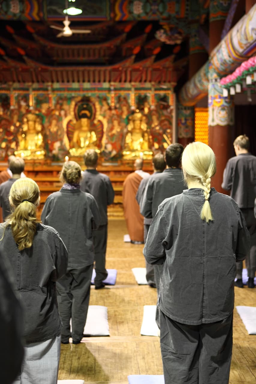 People in loose grey temple clothing standing quietly facing an altar inside a temple hall