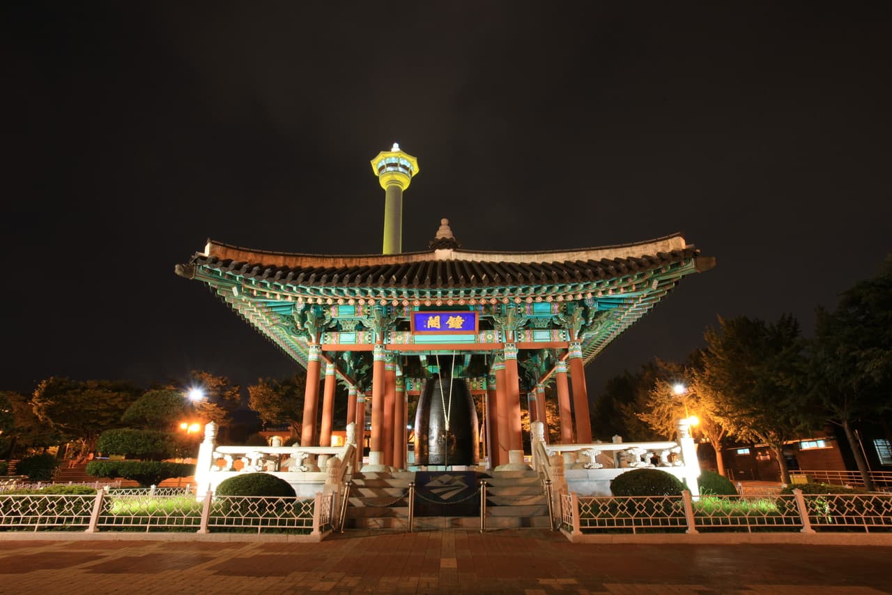 A traditional bell pavilion lit at night below Busan Tower in Yongdusan Park, Busan