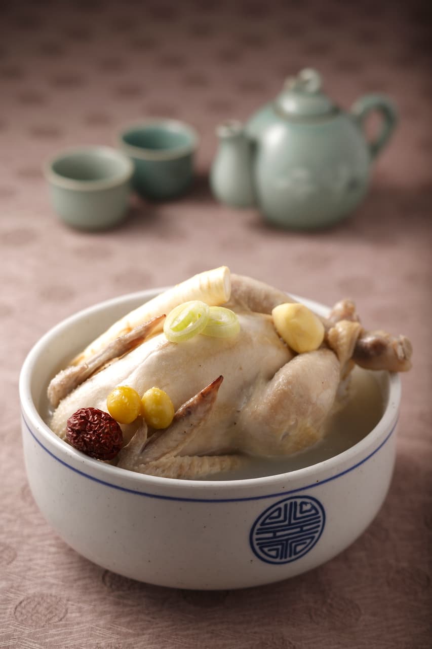 A whole young chicken in clear broth served in a white ceramic bowl with a celadon teaset behind