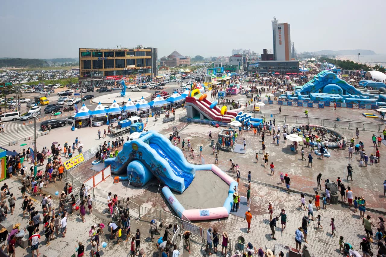 An aerial view of a beachside festival ground with inflatable mud slides, pools, and crowds