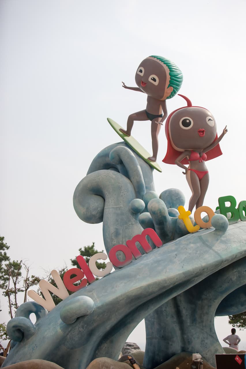 A playful mascot statue riding a wave sculpture above a "Welcome to Boryeong" sign