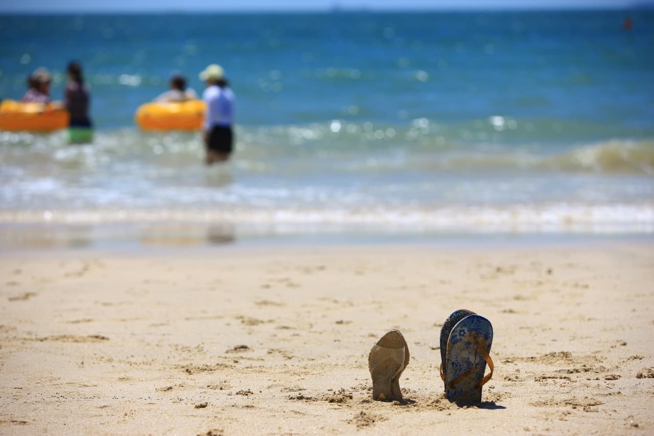 Sandals planted in beach sand with blurred swimmers and the sea behind
