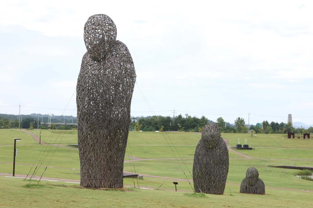 Large woven human-shaped sculptures standing on open green lawns in a public park