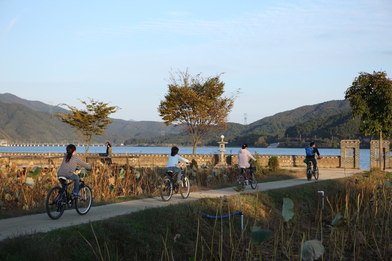 A riverside path with autumn trees and cyclists beside a broad river ringed by hills