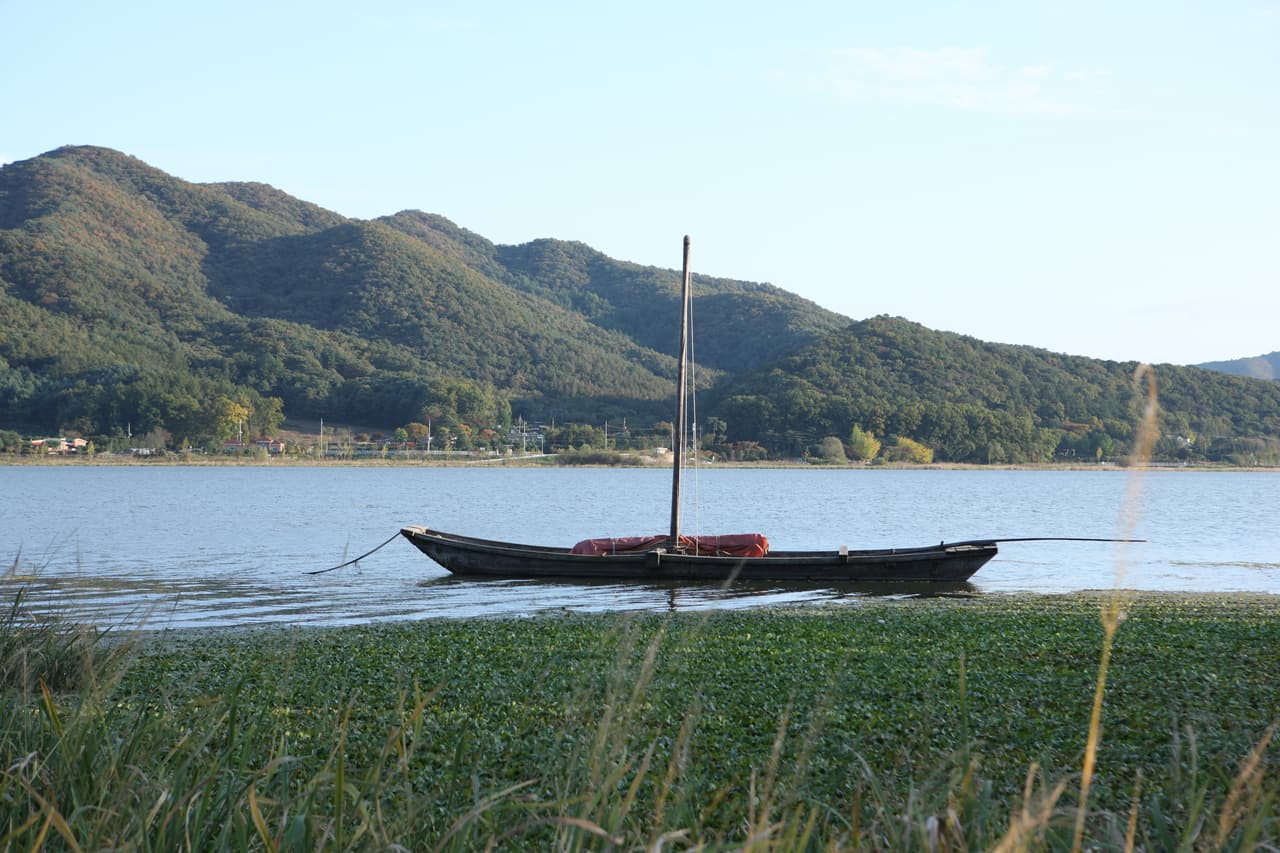 A traditional wooden sailboat moored on a wide calm river below wooded hills