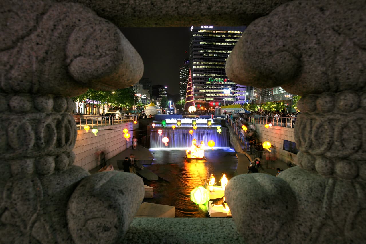 Glowing paper lanterns floating on a city stream at night beside a lit waterfall