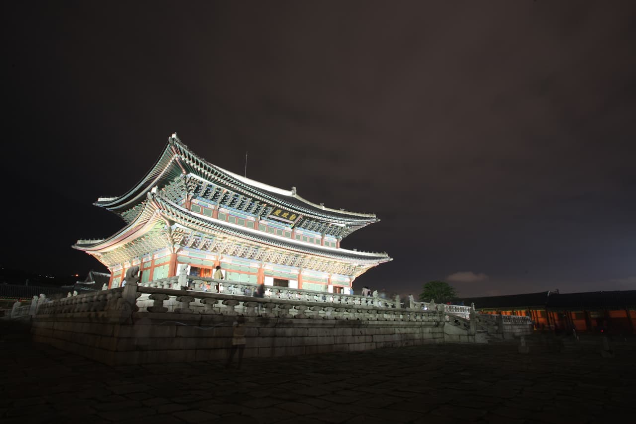 The lit main throne hall of a Korean palace at night, with stone terraces