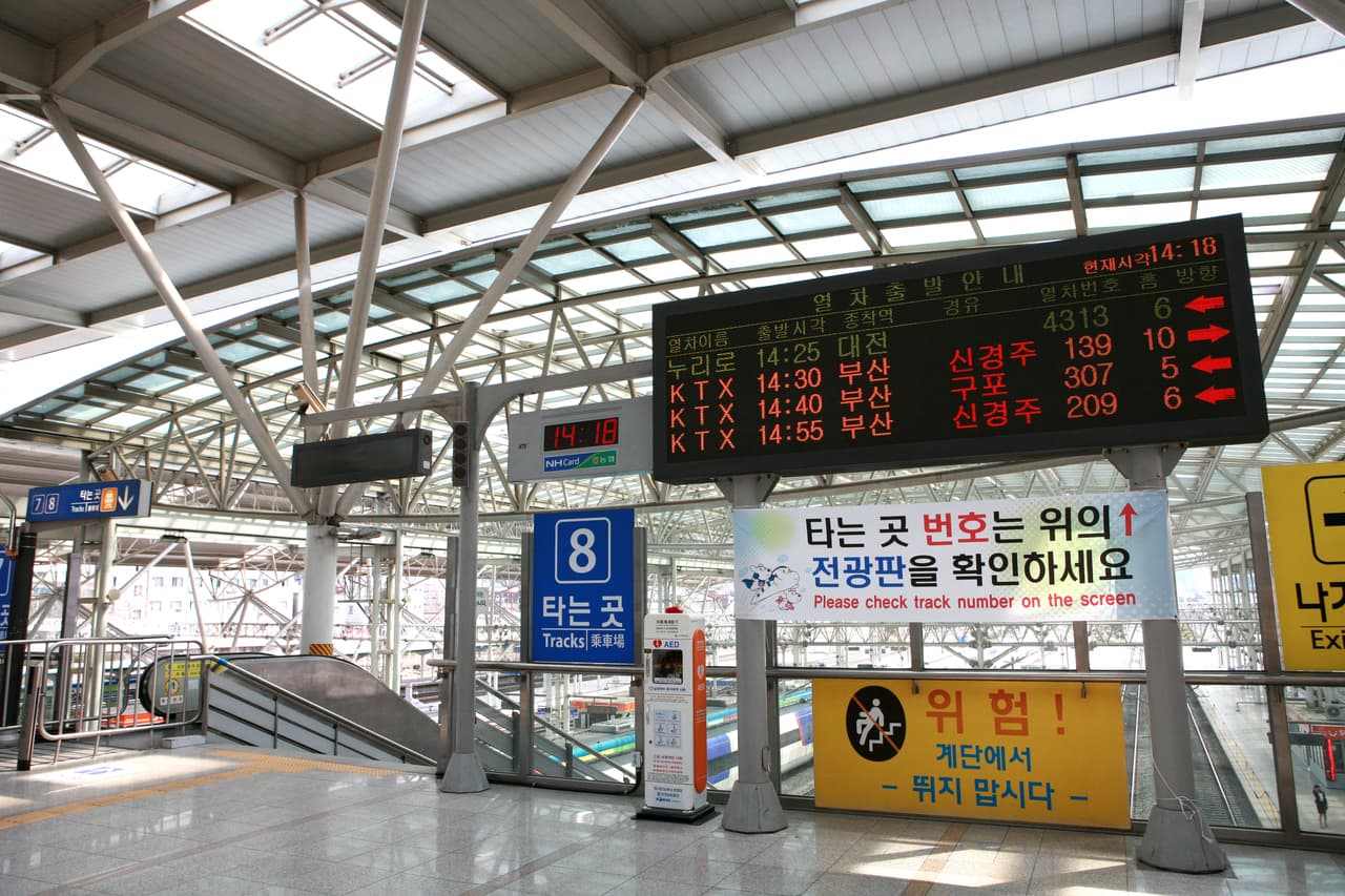 A station departure board and safety signs above a stairway on a train platform