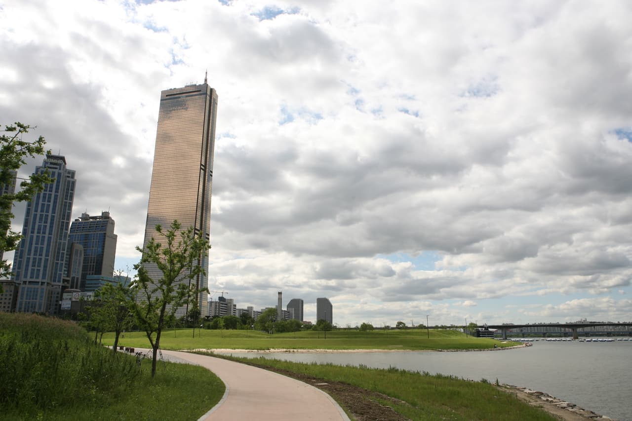 A green Han River park with a riverside path and the 63 Building under summer clouds, Seoul
