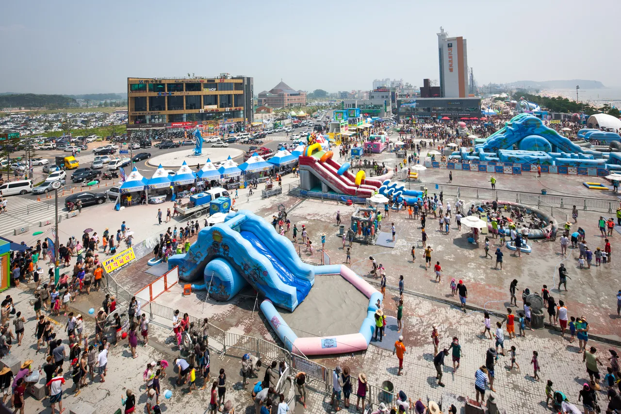 An aerial view of a beachside festival ground with inflatable mud slides, pools, and crowds