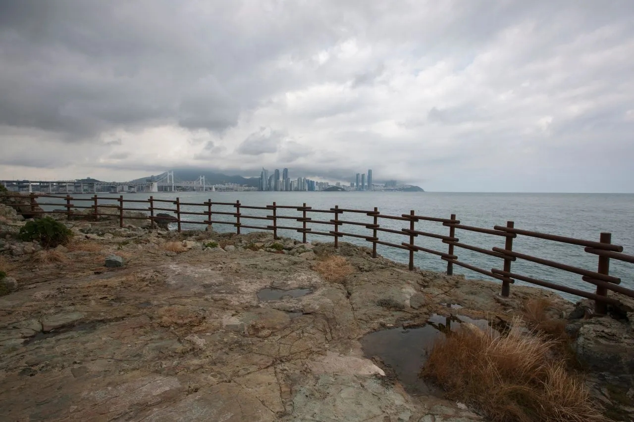 Rocky lookout on the Igidae coastal trail under a cloudy sky, with Gwangan Bridge and the Haeundae skyline across the water