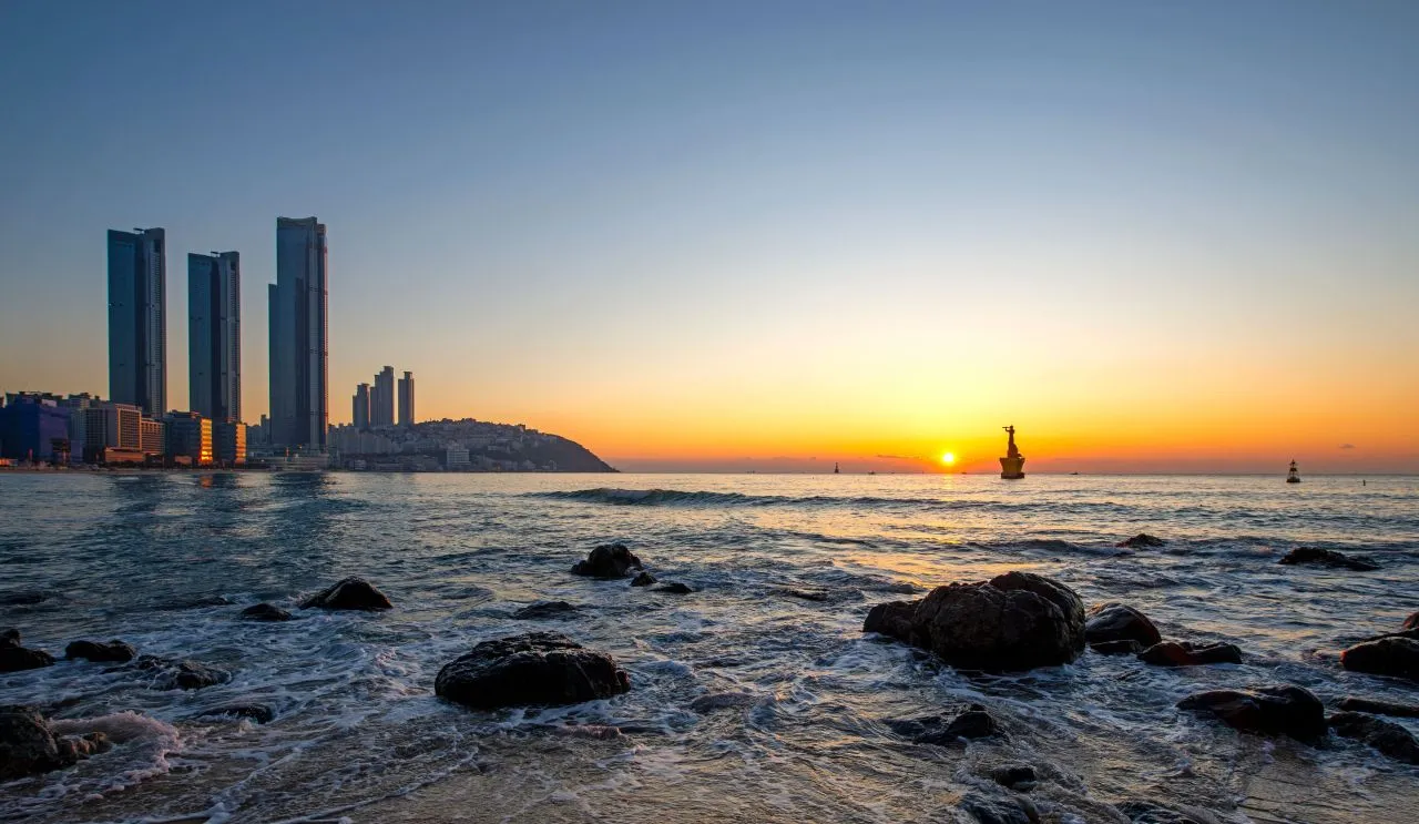 Low sun over the sea off Haeundae, with dark rocks in the surf and high-rise towers on the shoreline