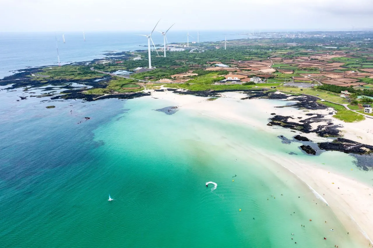Aerial view of a white sand beach with turquoise water, dark lava rock and wind turbines on the coast inland