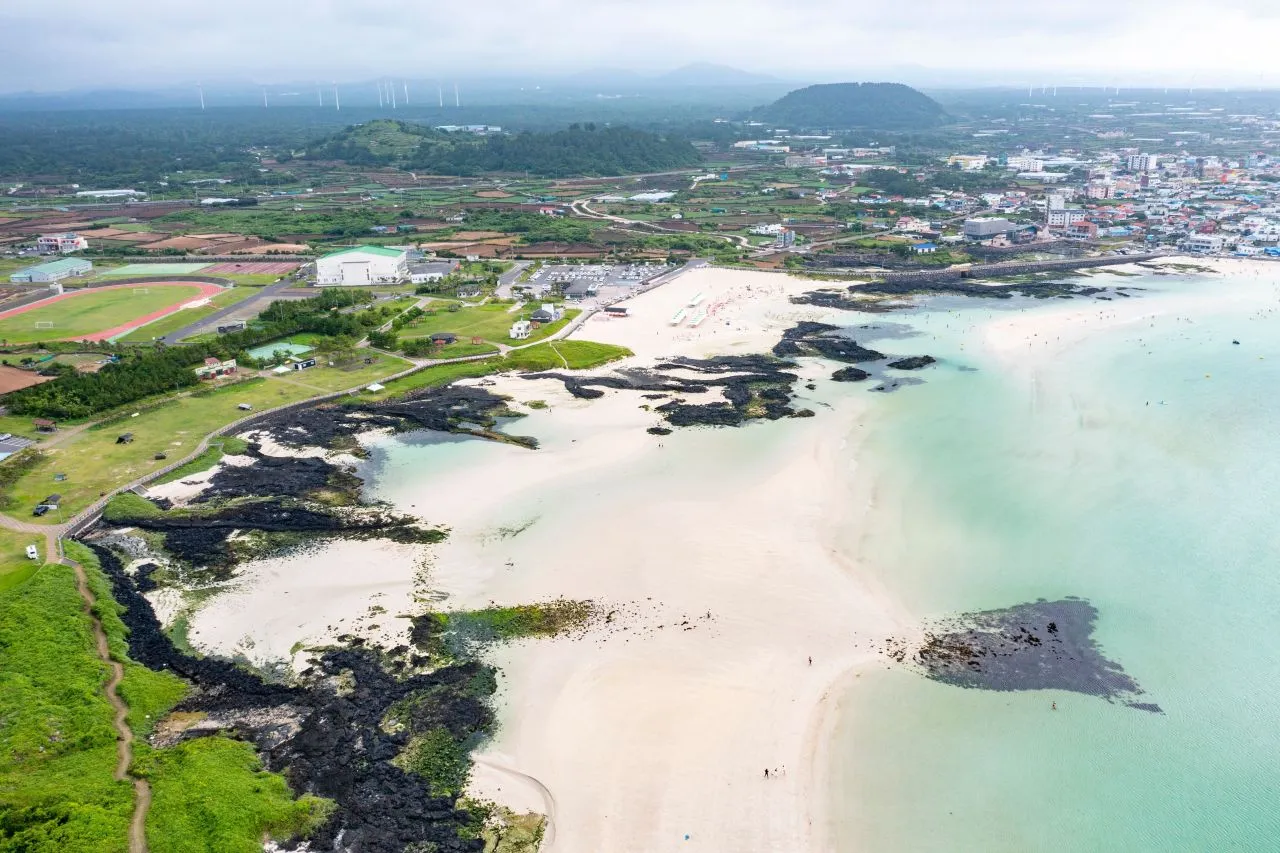 Aerial view of white sand, a dark lava rock outcrop and clear green water beside a coastal town