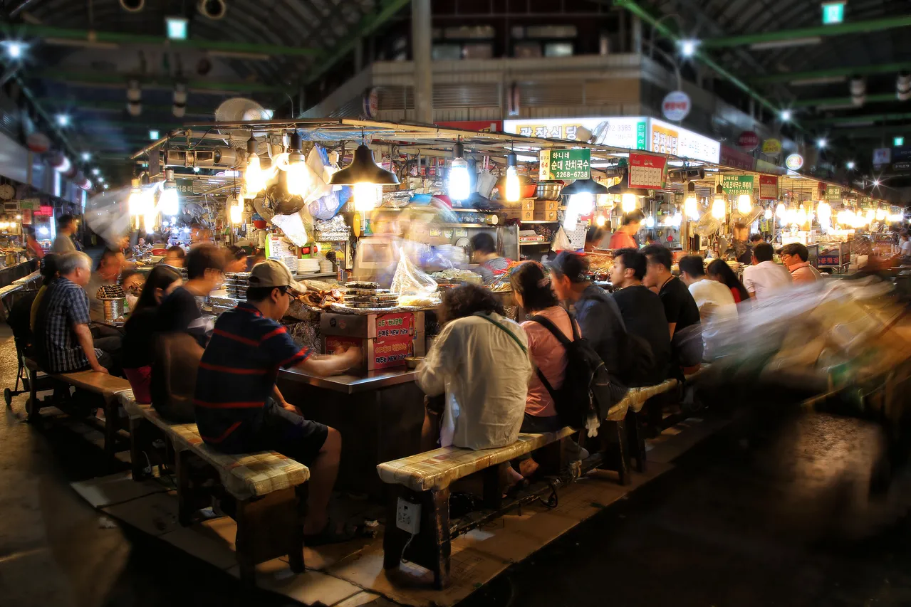 Diners seated at busy communal counters under lit stalls in a covered Seoul food market at night