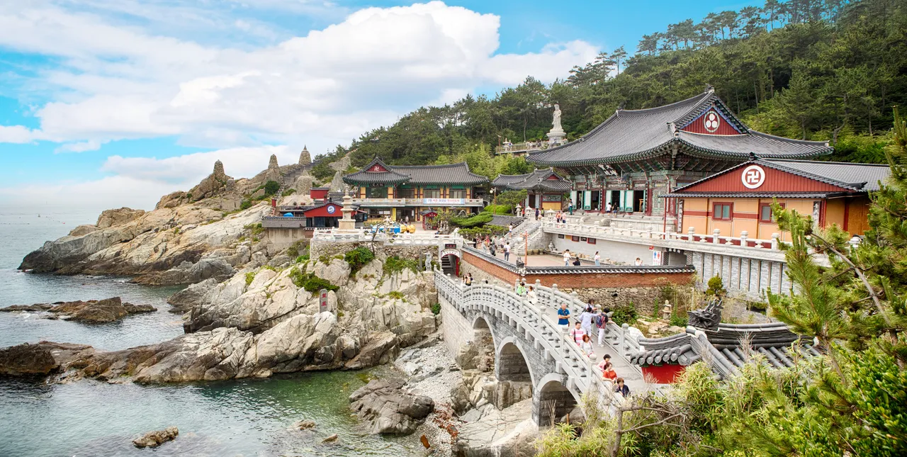Temple buildings on a rocky shoreline with an arched stone bridge in front and blue sea under scattered clouds
