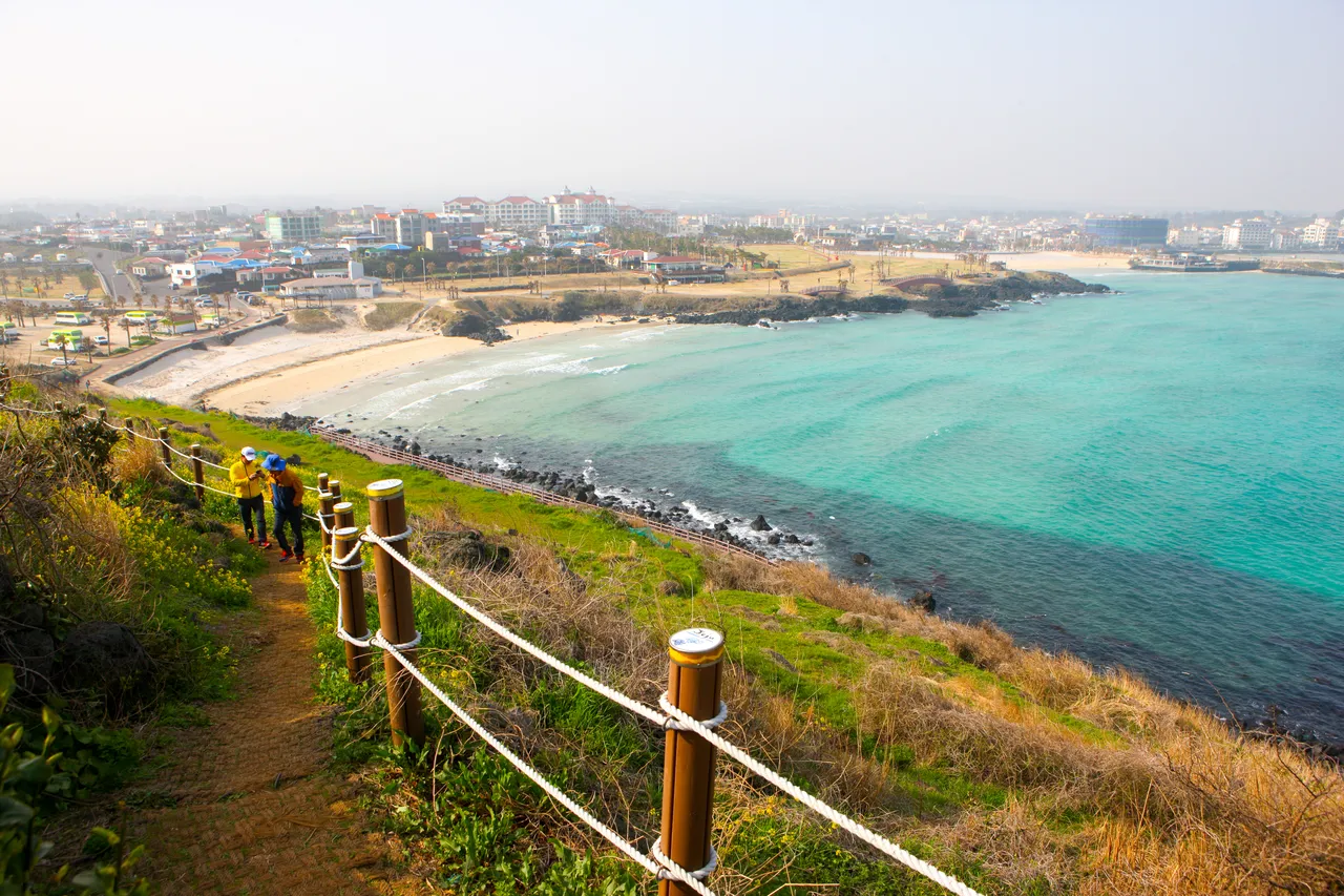 Hamdeok Beach seen from a hillside path with rope railings, with turquoise water curving along the sand