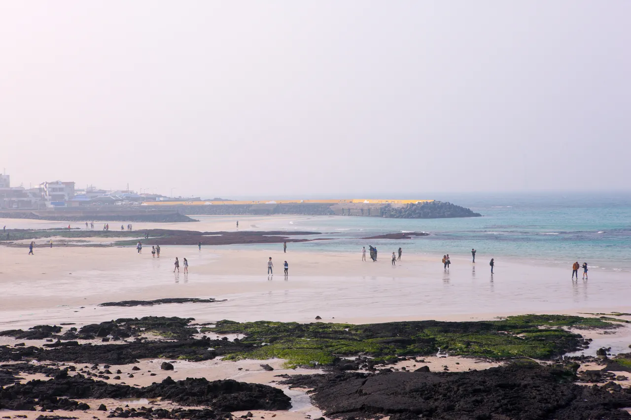 Wide sand beach with dark volcanic rock in the foreground and people walking toward the water