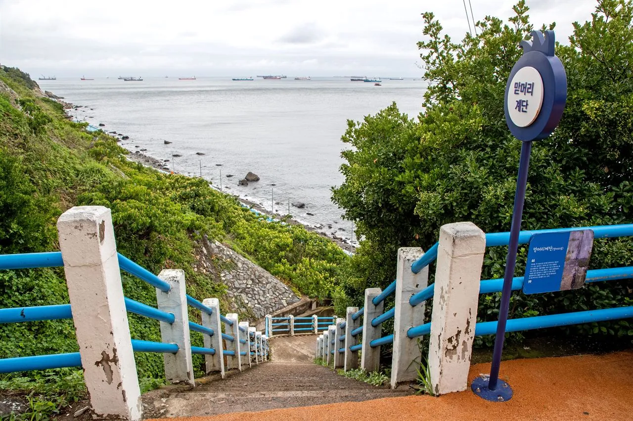 A stairway with blue railings leading down a green slope toward the sea, with a round blue sign beside it