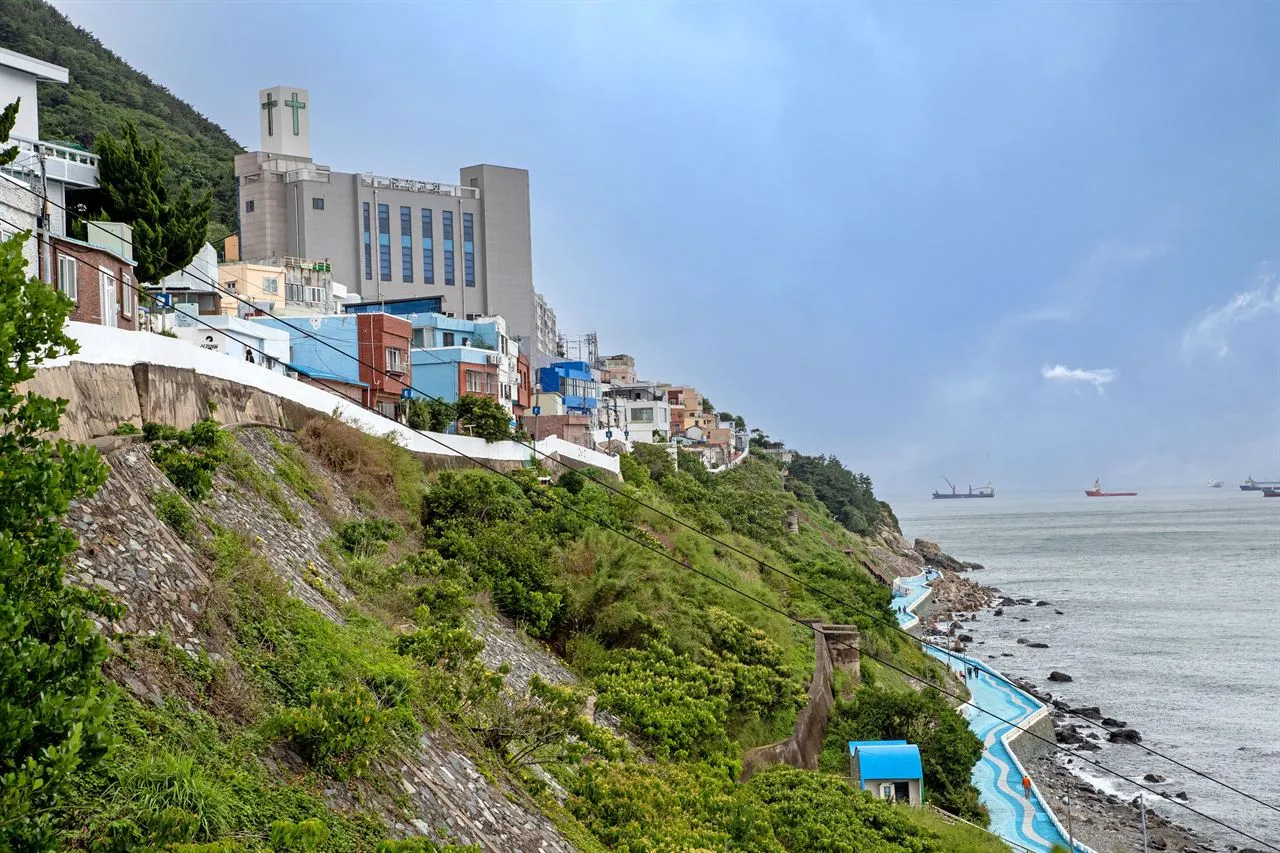 Painted and white houses stacked along a steep green hillside above a blue tiled coastal walkway and the sea