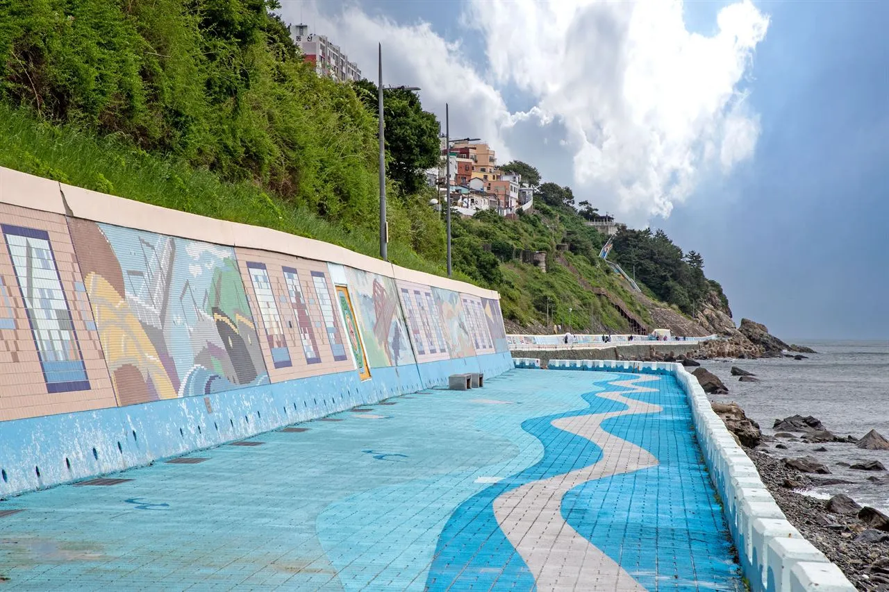 A blue tiled seaside walkway running beside a mosaic mural wall and a rocky shoreline
