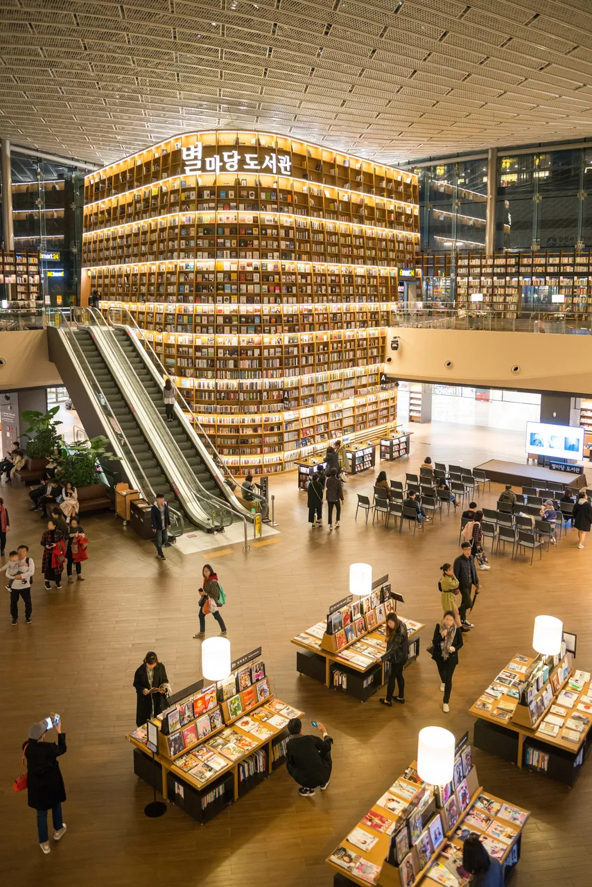 A lit wall of bookshelves rising several floors above an open indoor hall with escalators and book displays