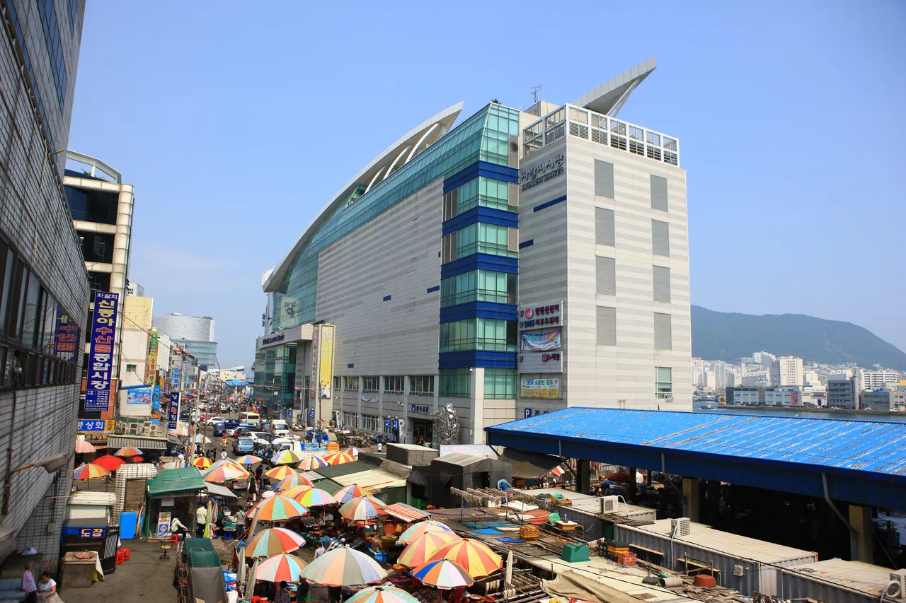 A curved-roof seaside market building behind rows of colorful parasols over street stalls, with a hill beyond