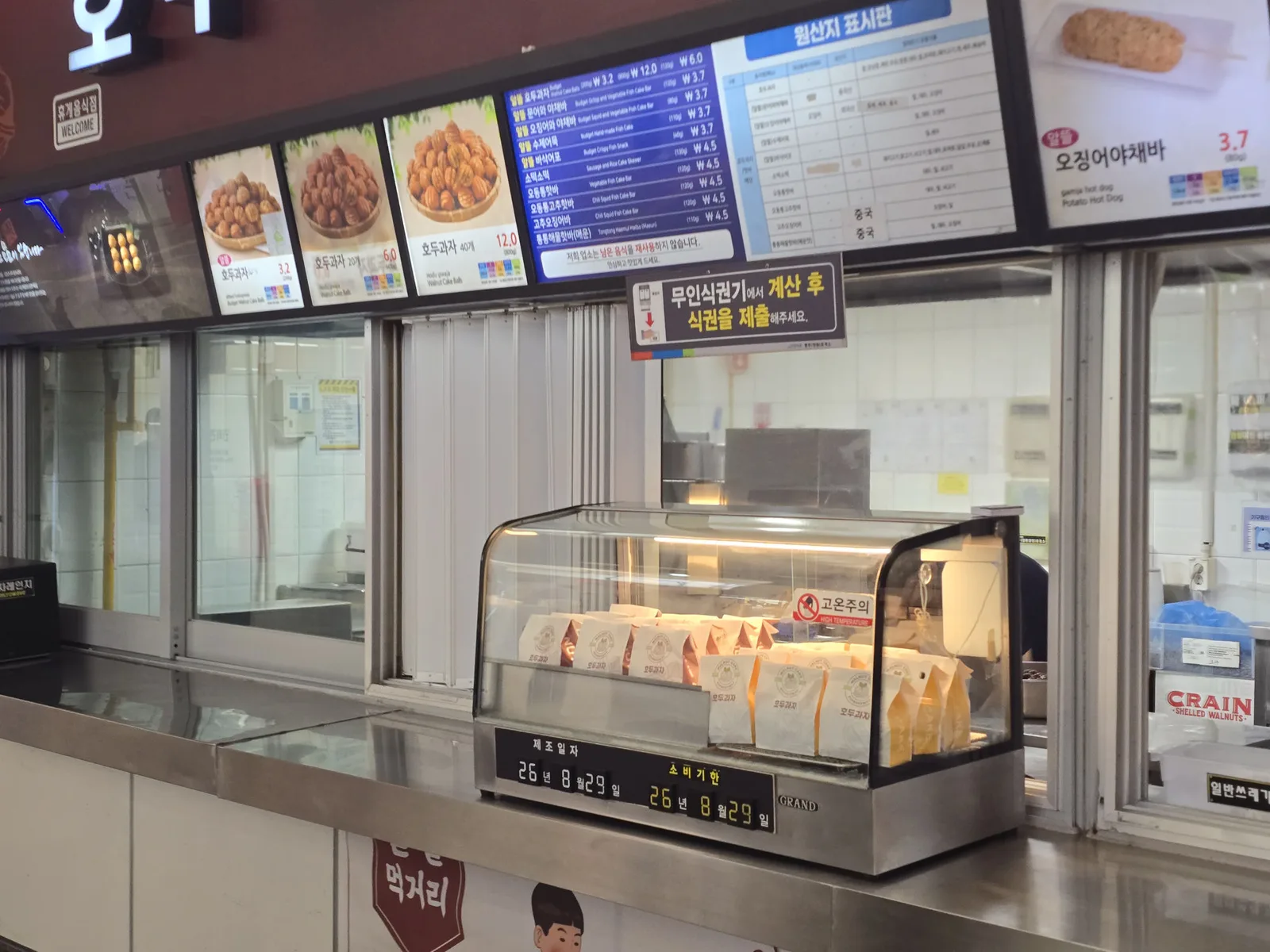 A rest area snack counter with walnut cakes in a warmer under menu boards and a notice about paying at a ticket machine