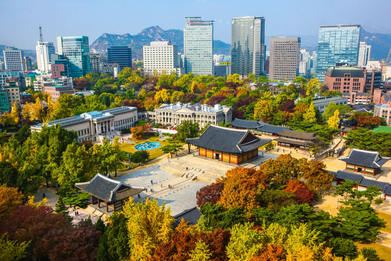 Yellow, orange and red autumn trees around Deoksugung palace halls in Seoul, with office towers and a mountain ridge behind