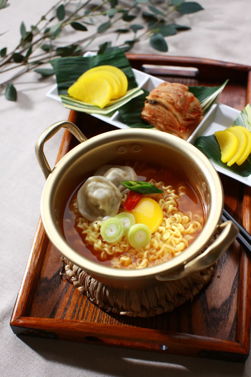A pot of Korean instant ramen cooked with an egg, dumpling, and scallions, with kimchi on the side