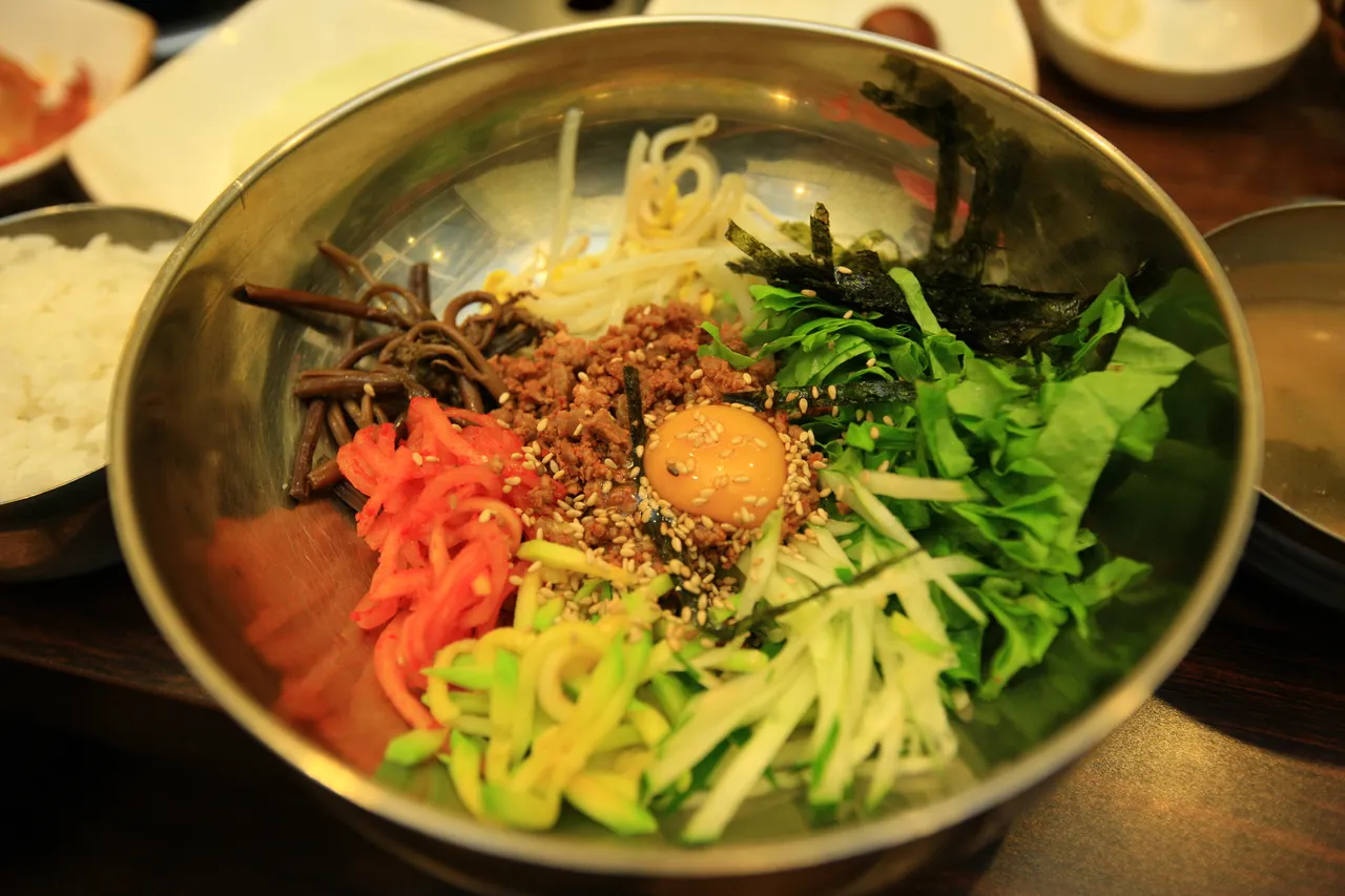 A metal bowl of bibimbap with seasoned vegetables, minced beef, and a raw egg yolk