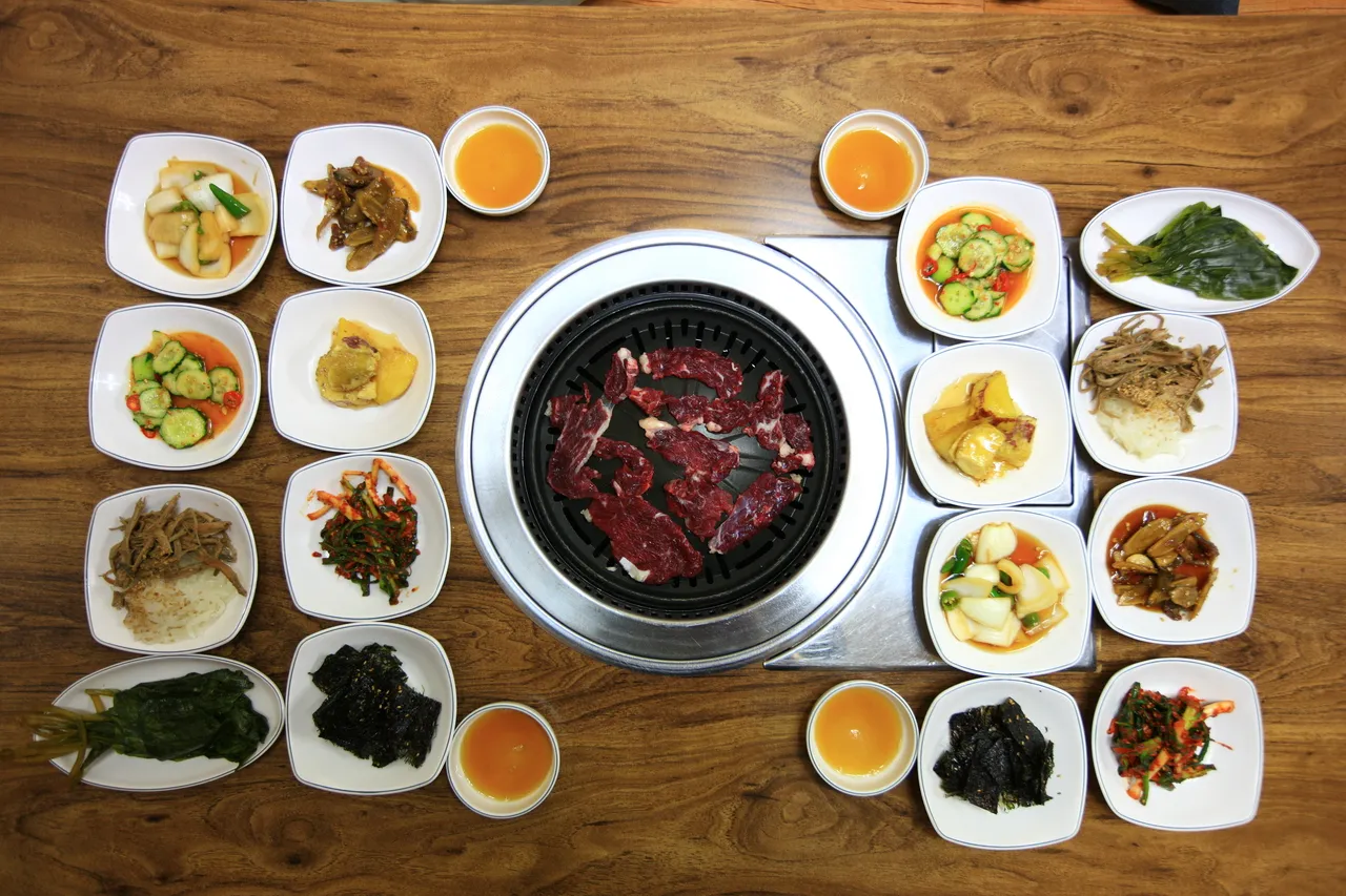 A Korean charcoal BBQ grill with raw beef in the center, surrounded by an array of banchan side dishes