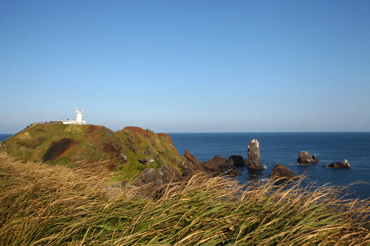 A white lighthouse on a green hilltop above the sea, with a rock stack standing offshore under a clear blue sky