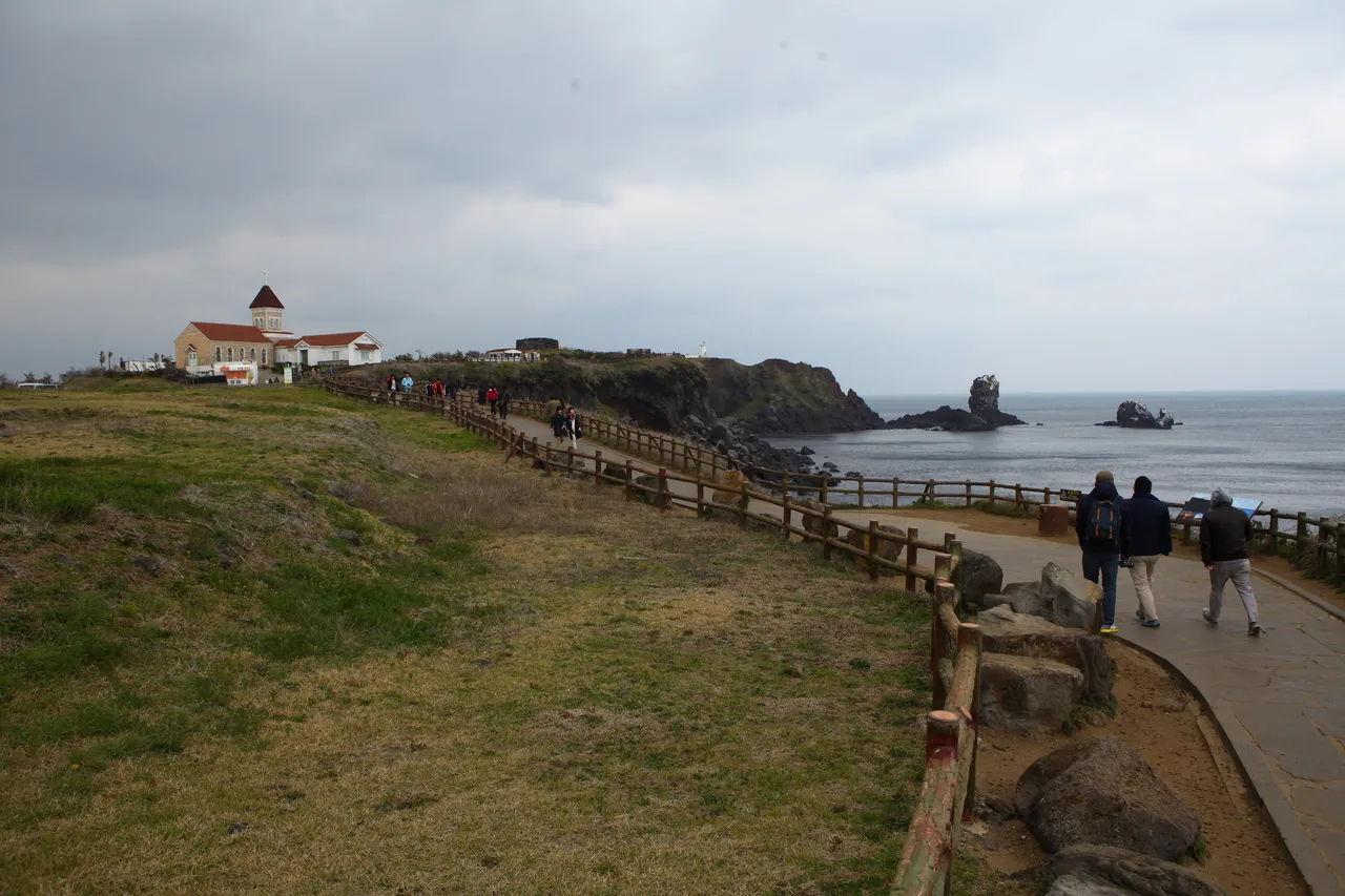 A railed coastal path crossing a grassy headland, with rock stacks in the sea and a cloudy sky beyond