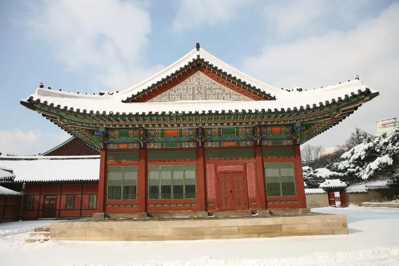 A red and green painted palace hall at Deoksugung in Seoul with snow on its tiled roof, snow-covered ground, and a snow-laden pine beside the wall