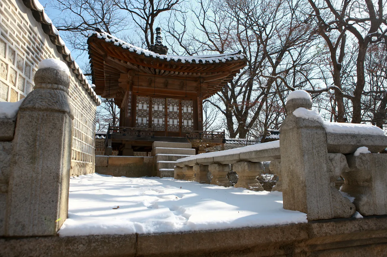 A small wooden pavilion with lattice doors in the Changdeokgung Huwon garden in Seoul, with snow on its roof, a stone balustrade, and bare trees