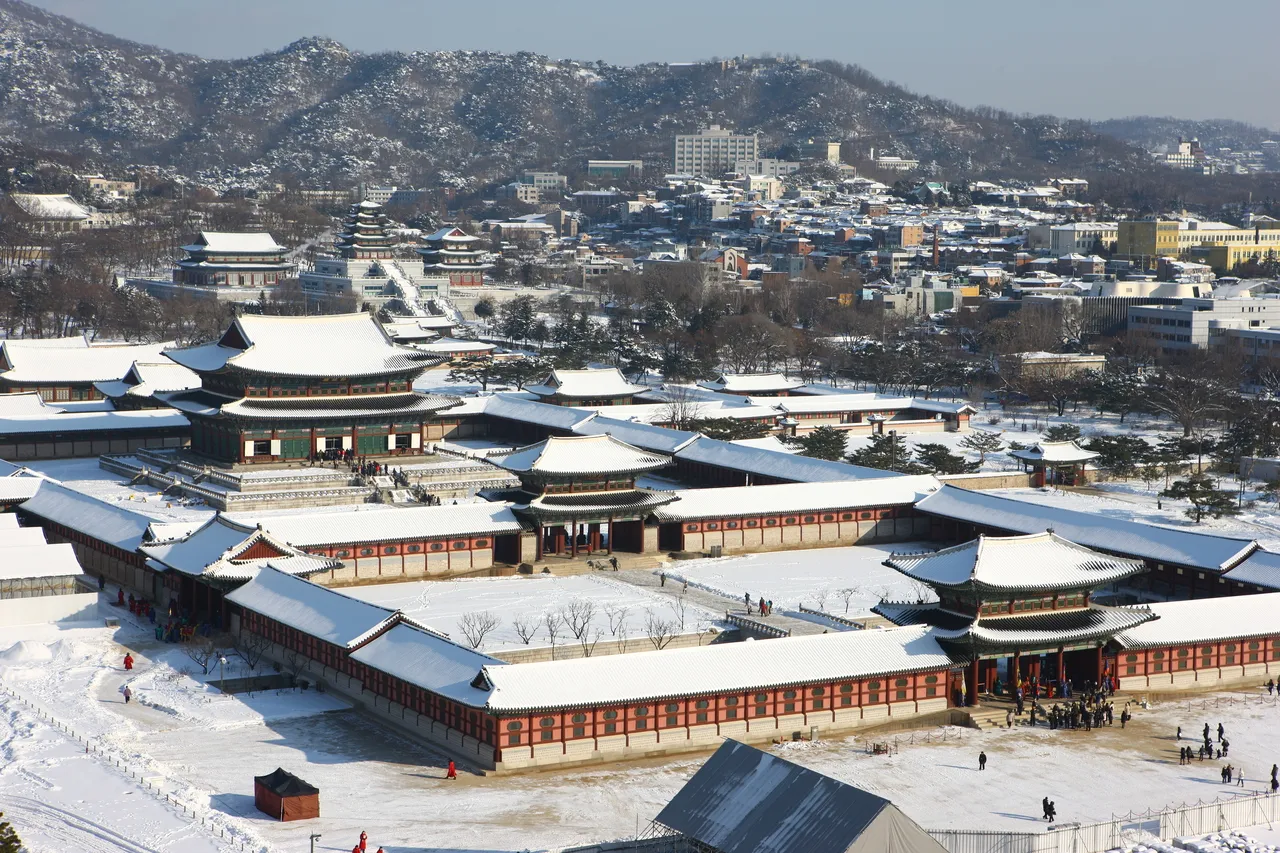 Gyeongbokgung palace halls in Seoul seen from above, with snow on the tiled roofs and courtyards and snowy hills behind