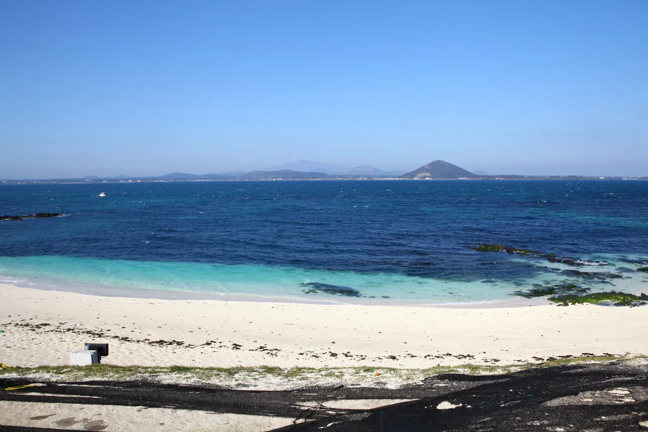 Pale rhodolith sand along the shore at Udo Sanho Beach with clear shallow water and rocks beyond