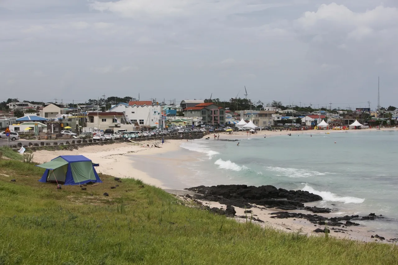 A sandy beach with small waves and dark rocks, with low buildings and parked cars along the road behind it