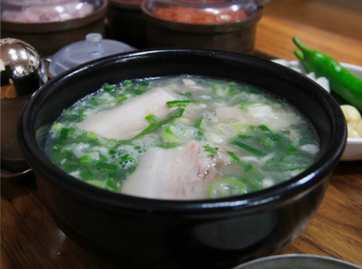 Close view of pork soup in a black stone bowl with sliced pork and chopped green onion, with seasoning pots and a salt shaker behind it