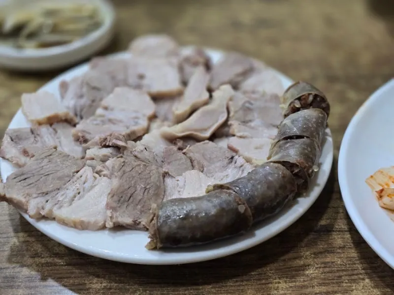 A white plate of sliced boiled pork with a coil of dark sundae sausage beside it