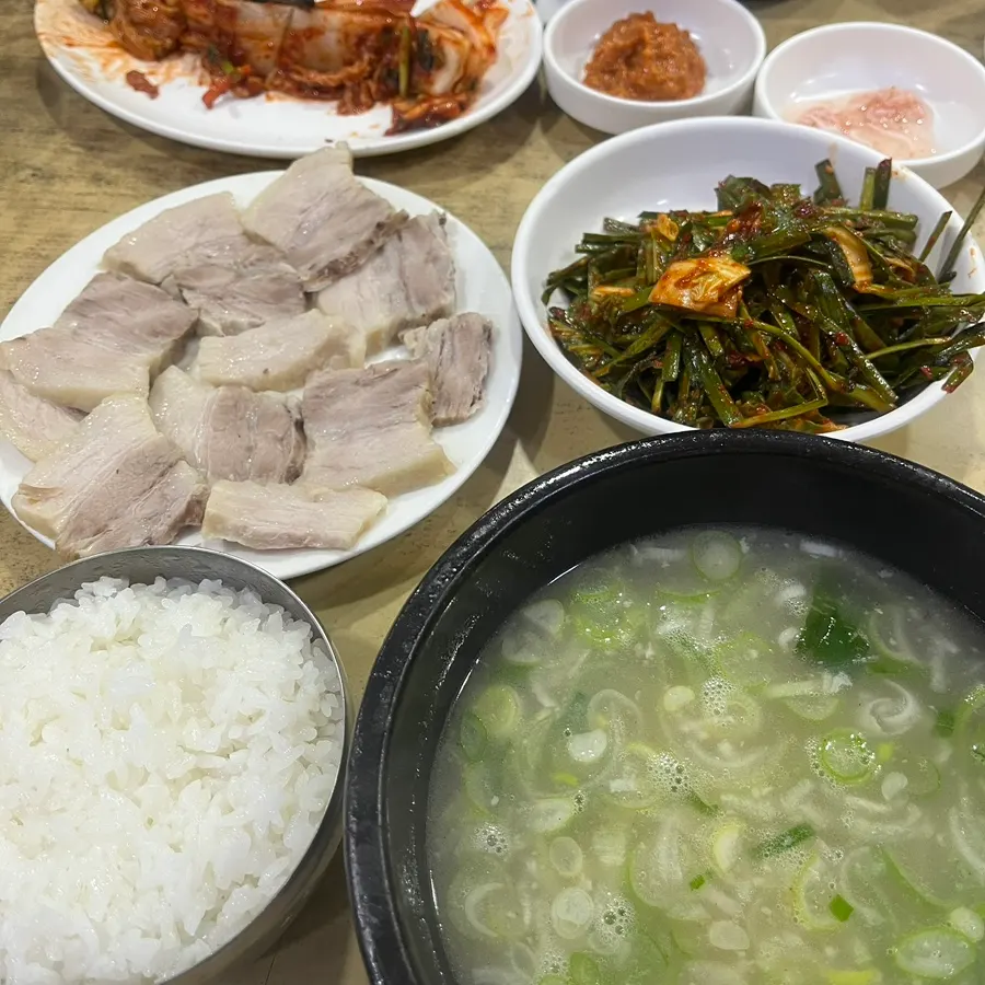 Boiled pork slices on a plate beside a bowl of rice, seasoned chives and a black bowl of clear pork soup